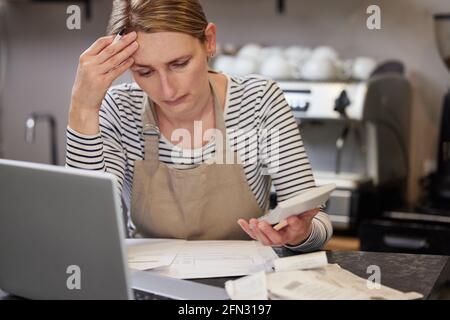 Worried Female Owner Of Coffee Shop In Financial Difficulty Looking Through Bills Using Laptop And Calculator Stock Photo