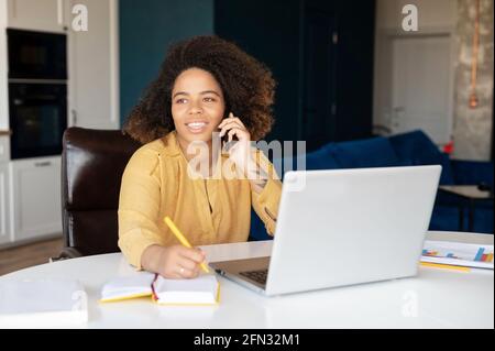 Cheerful African-American young woman talking on the smartphone, using laptop and taking notes at home, black female freelancer holding phone conversation and making schedule, writes down. Remote work Stock Photo