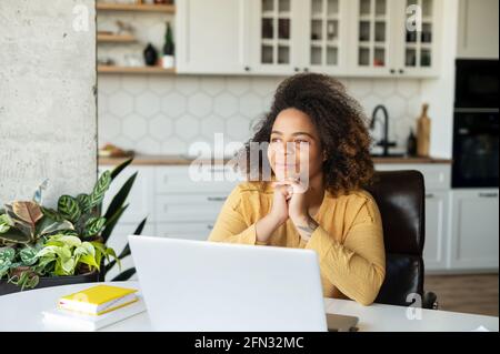 Delighted African-American young woman sits with a laptop in the kitchen in cozy apartment, looks away and dreaming, thoughtful happy dark skinned female freelancer lost in dreams and imaginations Stock Photo