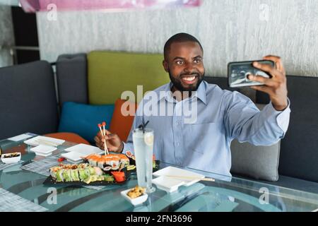 Cheerful young bearded African American man in light shirt doing selfie shot on mobile phone, posing at the table in cafe, eating sushi rolls served on black plate. Traditional japanese food Stock Photo