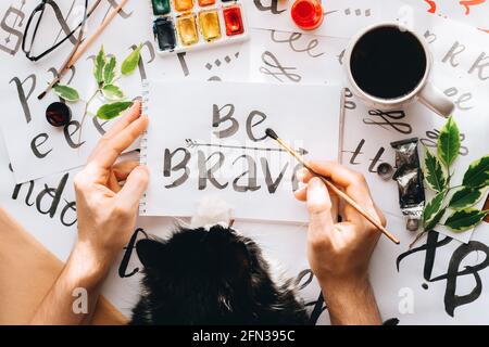 Top View man and cat writing on paper sheet canvas calligraphy word BE brave drawing with black red ink brush.creative artist freelancer in home studi Stock Photo