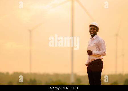 African engineer windmills wearing white hard hat and using tablet with   wind turbine on silhouette sunset Stock Photo