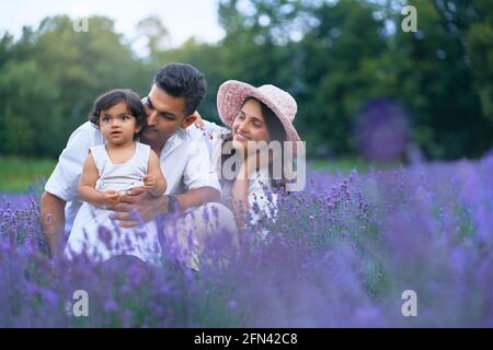 Front view of mother, father and daughter sitting in lavender field and smiling. Cute baby girl, carrying flower and enjoying time with loving parents outdoors. Young family, nature concept. Stock Photo