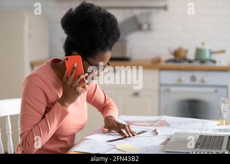 Concentrated black woman architect work on blueprint on phone call from home. Designer on freelance Stock Photo