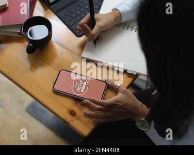 Overhead shot of female worker looking an idea on mock up smartphone and write it down Stock Photo