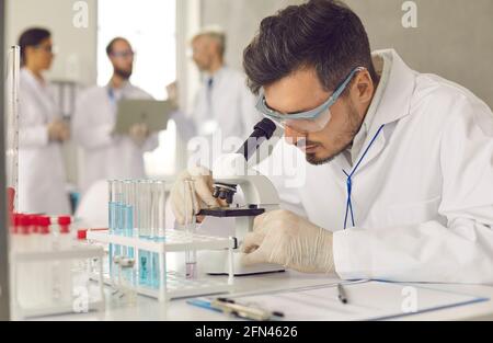 Serious scientist in lab coat, goggles and gloves looking at sample under microscope Stock Photo