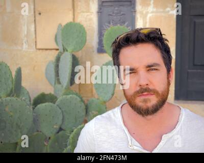Portrait of man with beard and narrowed eyes. Big prickly pear cactus in the background. Tourism and holidays in Italy. Stock Photo