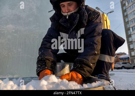 A worker assembler in winter overalls puts wet snow from a trough into a metal bucket Stock Photo