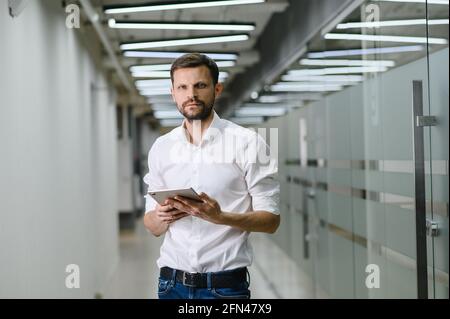 A man in a white shirt holds a tablet and smiles in the office. business man on the background of the office Stock Photo