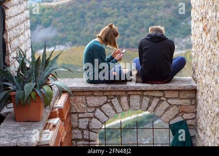 Teens seated on an arch. New technologies and ancient architectures. Teenagers use their mobile phones outdoors. Stock Photo