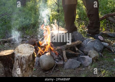 https://l450v.alamy.com/450v/2fn4bmp/campfire-above-the-flame-of-which-a-kettle-is-hanging-and-food-is-being-prepared-against-the-background-of-a-man-leg-in-the-camp-2fn4bmp.jpg