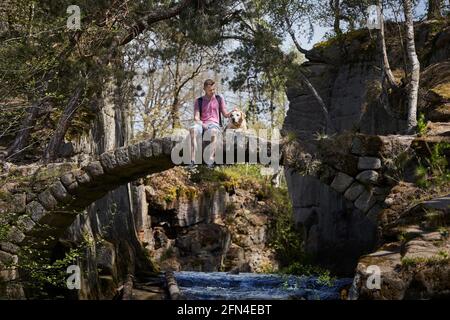 Young man with his dog sitting old stone bridge in beautiful nature. Tourist with labrador resting in the middle of the forest. Stock Photo