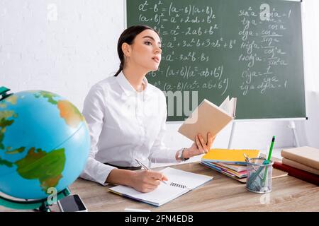 Teacher holding book and writing on notebook near globe in classroom Stock Photo