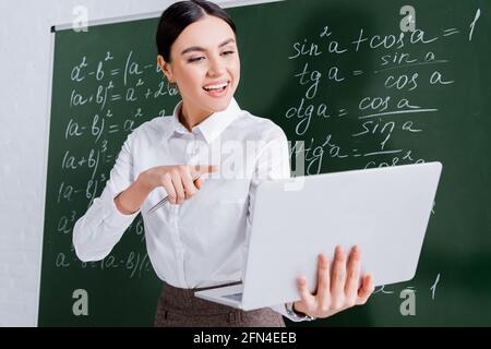 Smiling teacher pointing at laptop during video call near chalkboard in classroom Stock Photo