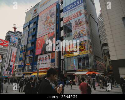 People walking in the streets of Akihabara. Businessmen and tourists stroll using smartphones in front of skyscrapers with huge manga billboards. Stock Photo