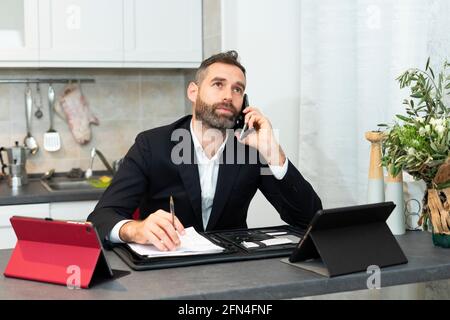 Busy young businessman calls and takes notes in the kitchen. Handsome man working at home. Smart working with tablets and sheets in covid pandemic. Stock Photo