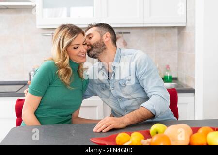 Happy couple is kissing in the kitchen. Newlyweds sitting at the kitchen table. Boyfriend kisses his blonde girlfriend. Good morning kiss. Stock Photo