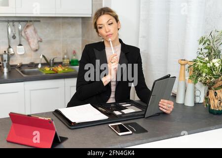 Attractive business woman thinks holding a tablet. A woman works from home on the kitchen table. Classy girl in suit wondering about what to write. Stock Photo