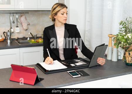 Attractive business woman works from home. Elegant girl in black suit using tablet and taking notes on the kitchen table. Smart working in lockdown. Stock Photo