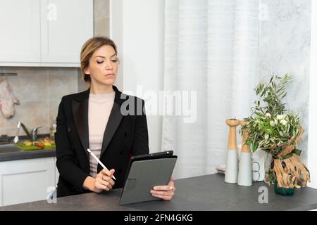 Attractive business woman works from home. Elegant girl in black suit using tablet and taking notes on the kitchen table. Smart working in lockdown. Stock Photo