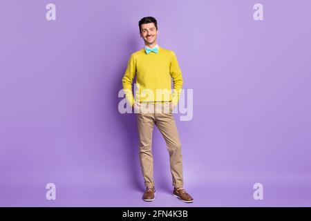 Full size photo of brunet optimistic guy stand hands in pockets wear yellow sweater trousers sneakers isolated on lilac background Stock Photo