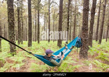 Mature male hiker using laptop in hammock in forest. Possible uses: Travel/remote working/adventure/ digital nomad/Shinrin Yoku concept image Stock Photo