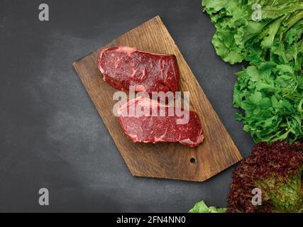 two raw pieces of beef steak on a brown wooden board, black table, next to green lettuce leaves, top view Stock Photo