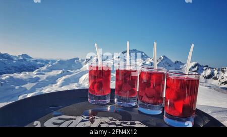 AL, GERMANY - Jan 02, 2020: Apres-ski. Four red schnapps with raspberries, on a brown tablet. In the background mountain panorama. Stock Photo