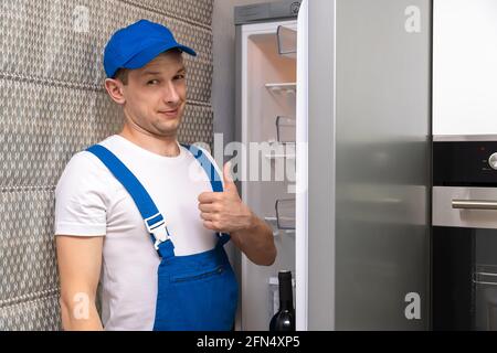 Uniformed repairman smiles, looks at the camera, and gives a thumbs-up sign against the backdrop of an open refrigerator in the kitchen Stock Photo
