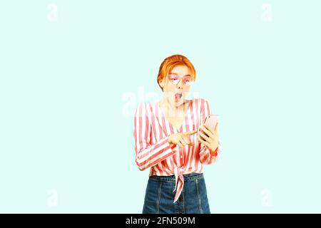 Portrait of a surprised natural redhead woman with open mouth wearing pink white stripes knot shirt & hipster cat eye sunglasses pointing finger at ce Stock Photo