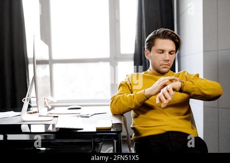 Close-up of a businessman wearing a yellow shirt sits near a large window in a modern office. Young freelancer looking at his wristwatch while waiting Stock Photo