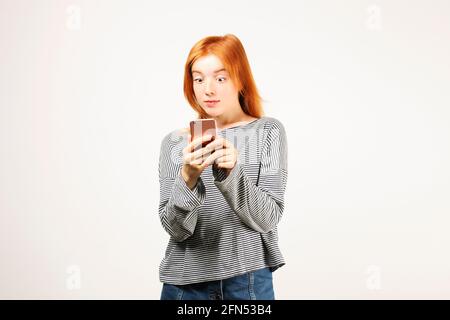 Portrait of surprised natural redhead woman wearing grey oversized loose shirt & hipster cat eye sunglasses staring at cell phone screen. Hipster fema Stock Photo