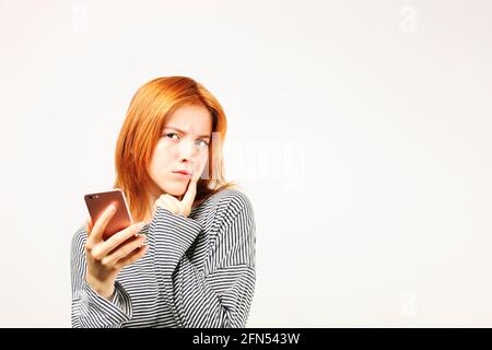 Young beautiful woman with long natural red hair, thinking facial expression and pose holding mobile cell phone. Attractive thoughtful hipster female Stock Photo