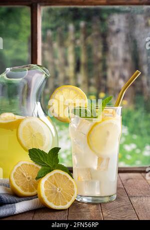 Glass of lemonade and pitcher with lemons on a wooden table by a window with rural background scene Stock Photo