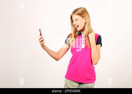 Lucky beautiful young happy woman with natural long blond hair celebrating her win, screaming and laughing, looking at her smartphone screen. Teenage Stock Photo