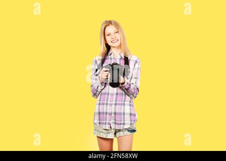 Young teenage girl holding digital photo camera with big lens & strap, taking pictures, smiling. Beautiful blond female photographer in checkered plai Stock Photo