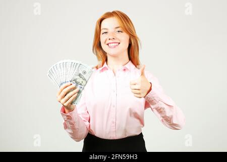 Young satisfied redhead woman showing thumbs up gesture, fistful of money holding one hundred dollar bill stack like fan. Excited attractive female w/ Stock Photo