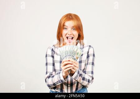 Young beautiful redhead woman screaming in rejoice, laughing w/ open mouth, fistful of one hundred dollar bills like fan. Excited attractive female wi Stock Photo