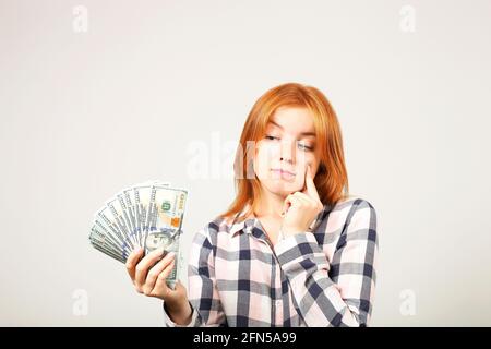 Close up portrait of young successful businesswoman holding bunch of hundred dollar bills, thinking where to spend her salary, touching chin with inde Stock Photo