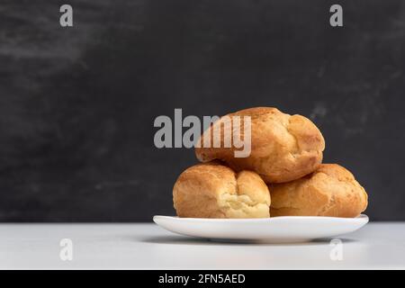 Three appetizing profiteroles on white plate. Homemade popovers and eclairs. Eclairs without cream. Stock Photo