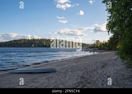 Upside down kayaks lie on the sandy shore, against the backdrop of the lake and rocky shore with trees. Beautiful scenery.  Stock Photo