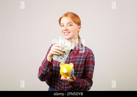 Portrait of young beautiful redhead woman in flannel shirt holding stack of hundred dollar bills in one hand & yellow piggy bank in another. Spend or Stock Photo