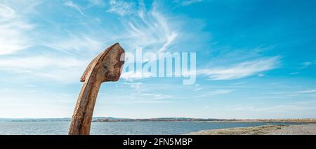 A rusty metal anchor against the river and the blue sky.  Stock Photo