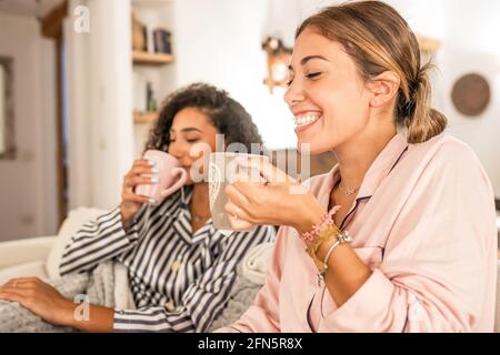 Couple of cohabiting girls in pajamas relaxing under blanket on cozy sofa drink a cup of tea while having fun talking and joking with each other. Real Stock Photo