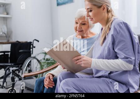 young social worker reading book to senior woman while sitting on sofa Stock Photo