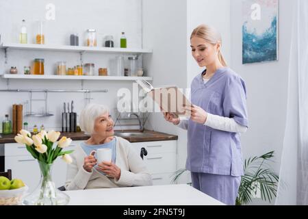 smiling social worker reading book to aged woman drinking tea in kitchen Stock Photo