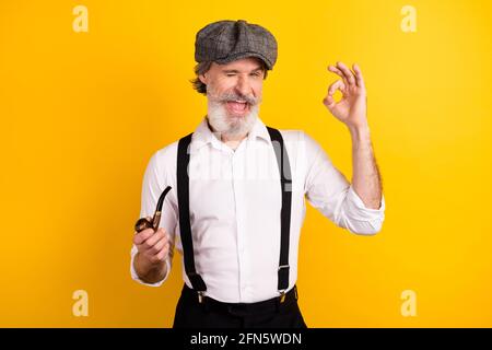 Photo portrait of gentleman winking keeping wooden pipe showing ok gesture isolated vibrant yellow color background Stock Photo