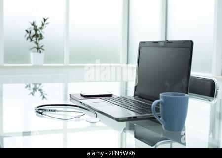 Laptop medical stethoscope coffee cup phone day planner on glass table in doctor's bright office space Stock Photo