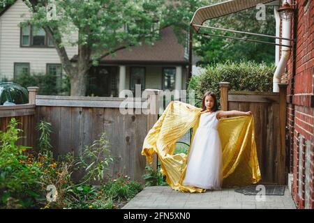 A little girl stands in garden arms outstretched in long gold cape Stock Photo