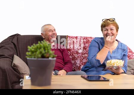 Senior couple watching tv on the sofa near table with digital tablet at home, smiling eating popcorn. Strong and harmonious family relationships Stock Photo